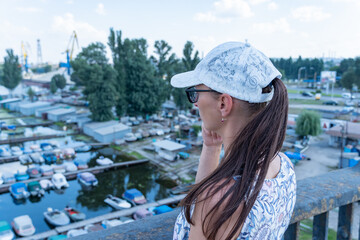 Woman in white cap thoughtfully looks at picturesque river and motor boats parked. She stands on...