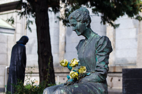 La statua bronzea di una donna seduta con delle rose gialle in grembo su una tomba del cimitero maggiore di Milano