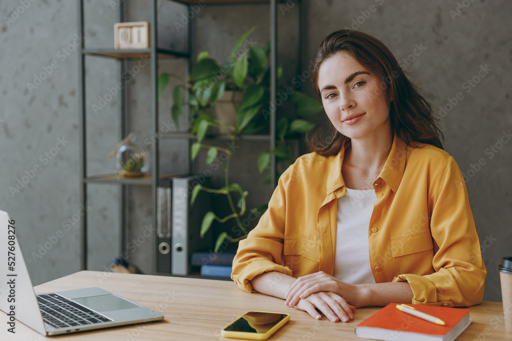 Wall mural Young successful calm cheerful confident employee business european woman 20s wear casual yellow shirt look camera sit work at wooden office desk with pc laptop. Achievement career occupation concept.