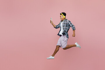 Fototapeta na wymiar Full body side view young excited man of African American ethnicity 20s wear blue shirt use mobile cell phone listen music in headphones isolated on plain pastel light pink background studio portrait.