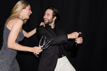 Fototapeta na wymiar Cheerful woman in dress holding glasses while boyfriend opening champagne isolated on black.