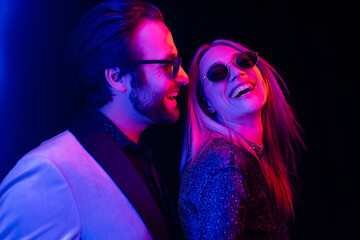 Fashionable man looking at girlfriend in sunglasses isolated on black with colorful lighting.