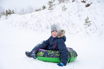 Fototapeta na wymiar Happy smiling little boy slide down the ice slide, sits on an inflatable tubing. Cute little happy child having fun outdoors in winter on sledge. family winter time.