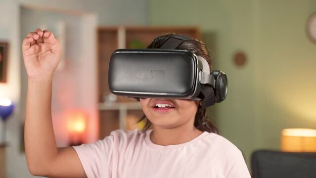 Smiling girl kid with VR or virtual reality goggles watching videos or experiencing virtual world on metaverse by touching objects at home - concept of immersive experience and modern technology.