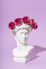Card with an artificial statue of David in a wreath of flowers on a purple background. Creative modern greeting card