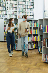 teenage students in medical masks walking with notebooks and laptop in library.