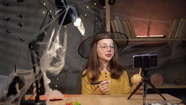 Teen blogger witch recording video blog. speaking looking at smartphone on tripod at home table on Halloween. girl social media vlogger shooting vlog about Halloween craft
