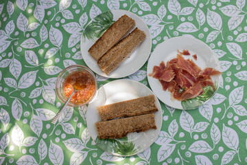Close-up of a small portion of toasted bread, serrano ham and tomato, and oil ready to eat on a table with floral motifs outside a garden. Food and vacations.