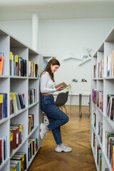 side view of teen girl standing near racks in library and reading book.