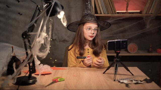 Recording Review. Cheerful witch teen girl filming her blog talking to camera of smartphone on tripod, sitting at table on Halloween. showing pumpkin on camera