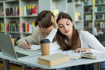 teenage girl reading book near friend writing in notebook and laptop in library.