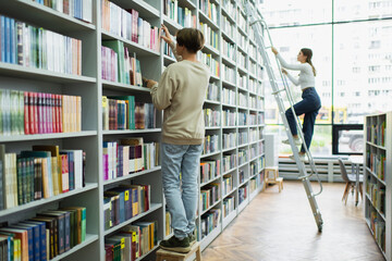 full length of teenage students choosing books on racks in library. - Powered by Adobe