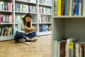 teen girl sitting on floor with crossed legs and reading book in library on blurred foreground.
