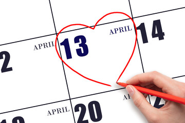 A woman's hand drawing a red heart shape on the calendar date of 13 April. Heart as a symbol of...
