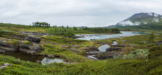 Panoramic view of blue glacial Arasjahka river rapids in Lapland landscape with green mountains and birch trees at Padjelantaleden hiking trail, north Sweden wild nature. Summer cloudy day