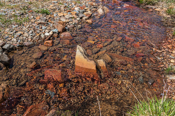 Sharp stone in clear water stream with motley stony bottom in sunlight. Colorful mountain stream with mineral water in bright sun. Picturesque top view to sunlit multicolor stones in spring water.