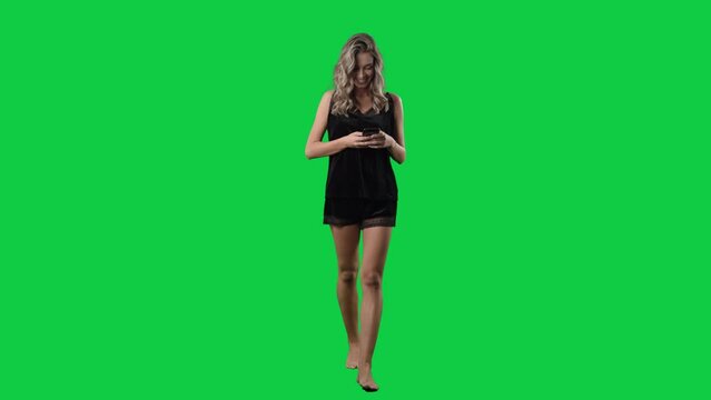 Happy young woman in black sleepwear using cell phone browsing internet. Full body isolated on green screen background