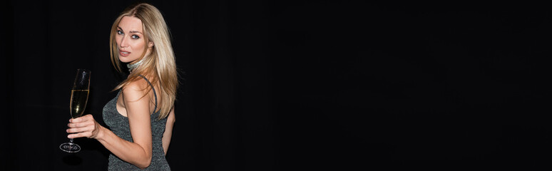 Trendy woman in dress holding champagne and looking at camera isolated on black, banner.