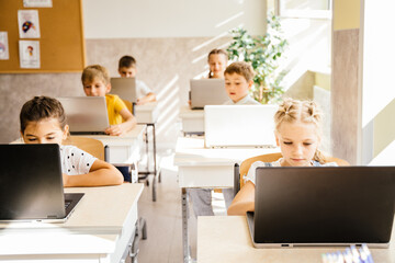 Education and school concept. Elementary school computer science class. Junior students girls and boys sitting at desk use laptops at lesson. Modern education concept.