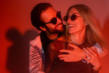 Young man in sunglasses embracing stylish girlfriend during party on red background.