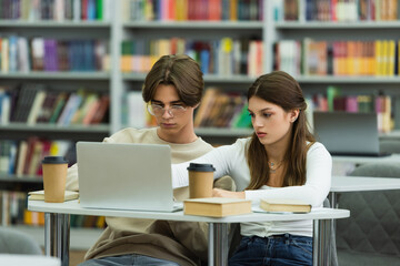 teenage girl and guy in eyeglasses studying near laptop and books in library.