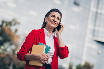 Photo of dreamy positive senior boss woman dressed red blazer communicating modern device outdoors urban town street