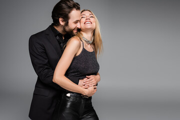 Bearded man in jacket hugging smiling blonde girlfriend isolated on grey.