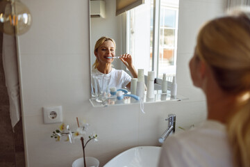 View over shoulder on young pretty blonde female brushing teeth at mirror in bathroom.