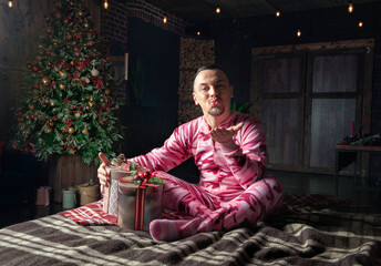 Cheerful funny man in pink sleepwear sit on the bed near decorated fir tree and send kiss