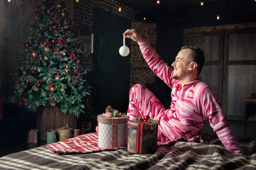 Cheerful funny man in pink sleepwear sit on the bed near decorated fir tree with Christmas present