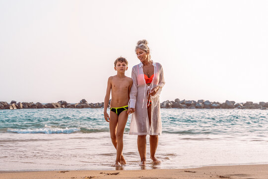 portrait of beautiful middle aged woman with her teen son in swimwear walking on the beach - Powered by Adobe