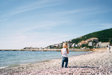 Little girl stands on a pebble beach and looks at the mountains. High quality photo