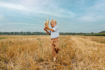 Naklejka na ściany i meble The blond boy is having fun playing with a straw hat in the summer in the field. The boy throws his hat into the sky. A happy child. Summer. Rural area