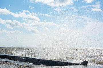 The waves break on the pier and there are a lot of splashes and water in the air, a windy and sunny...