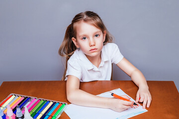A little girl in a white polo t-shirt with beautiful big eyes sits at the table and draws with a...