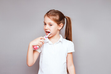 Fototapeta premium A little girl with two tails in a white polo t-shirt on a gray background brushes her teeth with toothpaste with an electric brush. Oral dental care concept, advice from a dentist