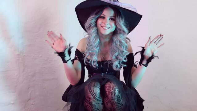 Young beautiful witch in halloween costume, wearing a witch's hat, standing around halloween decorations with lights, sits down and waves smilingly