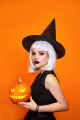 Beautiful sexy woman in witch hat and costume holding carved pumpkin on orange background.