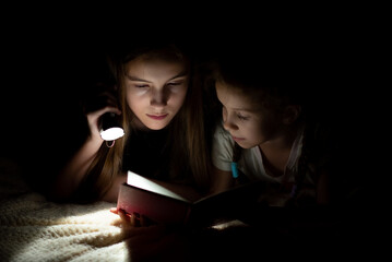 A teenage girl with her sister read in the dark with a flashlight.