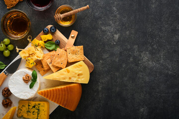 Obraz na płótnie Canvas Assortment of cheese, honey, cracker, blueberries, grapes with red and white wine in glasses antipasto server on white marble board on old dark grey background. Flat lay, copy space.
