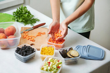 Woman holding slice of carrot above plastic container at table in kitchen. Preparing raw vegetables for freezing for winter storage in trays.