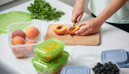 Hands of young female chopping fresh peaches on wooden board while preparing fruits and vegetables for freezing. Stack of plastic containers with raw cut vegetables for freezing on kitchen table.
