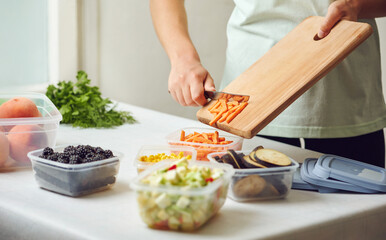 Hands of young female holding knife and wooden board with fresh slices carrot while preparing fruits and vegetables for freezing