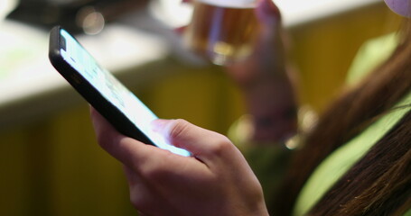 Fototapeta na wymiar Closeup of young woman hand using cellphone touchscreen while drinking alcholic beverage at bar nightlife