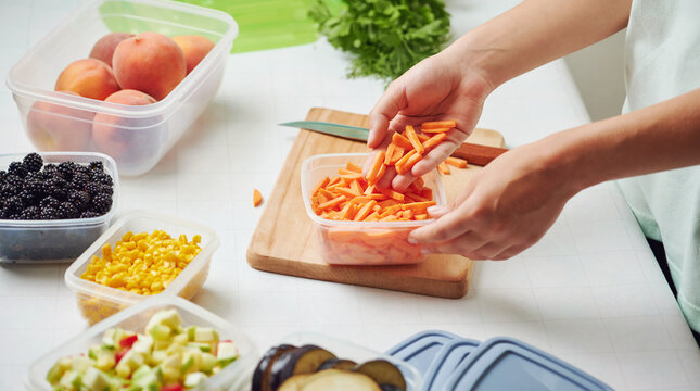 Woman holding plastic container with slices of carrot at table in kitchen. Raw vegetables for freezing for winter storage in trays.