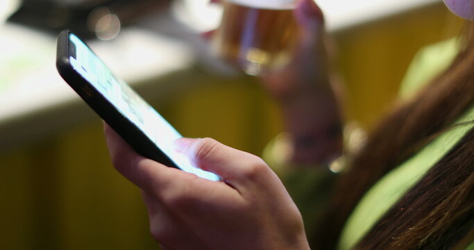 Closeup of young woman hand using cellphone touchscreen while drinking alcholic beverage at bar nightlife