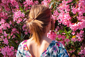 Naklejka premium Young woman stands with her back in beautiful pink flowers. Girl in a colorful linen dress between flowers. Back view. Vertical photo