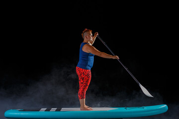 Fototapeta na wymiar A woman with a mohawk on a sup board kneeling with an oar on a black background.