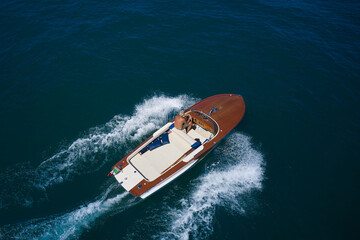 Italian wooden speedboat moving up aerial view. Wooden expensive boat fast movement on dark water top view. Big expensive wooden open boat with a man moving fast on dark blue water top view. - obrazy, fototapety, plakaty