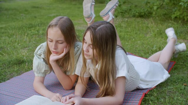 Teenage girls reading book together while lying on grass. Dolly shot of females spending leisure time at park. They are wearing casuals during summer. High quality 4k footage